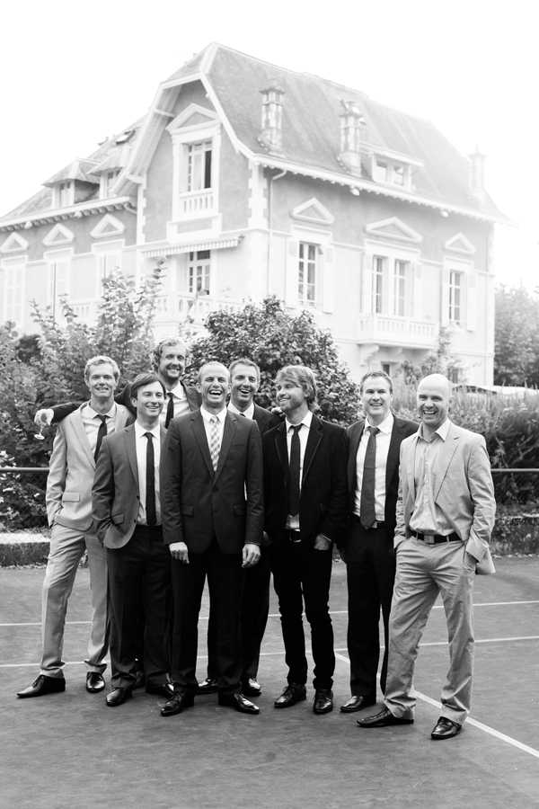 Black-and-white portrait of a group of eight men, likely the groom and his groomsmen or male guests, posing and laughing together outdoors on what appears to be a tennis court. The men are dressed in a mix of dark and light suits with ties, in varying styles of smart-casual to formal attire. The image is taken from a medium distance and captures the group in a candid, relaxed moment with genuine laughter. In the background stands a large French manor-style villa with a steeply pitched roof, dormer windows, and decorative architectural detailing, slightly overexposed against a bright sky.