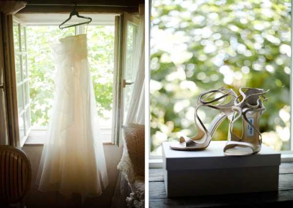 A split detail shot showing two bridal accessories during the getting-ready phase. On the left, an ivory strapless ballgown with a layered tulle skirt hangs from a hanger in an open window, backlit by natural light filtering through green foliage outside. On the right, a pair of nude strappy high-heeled sandals, identifiable as Jimmy Choo by the visible label, are placed on top of a white shoe box on a dark surface, with soft natural light and bokeh foliage in the background. Both images are close-up detail shots with a soft, naturally lit aesthetic.