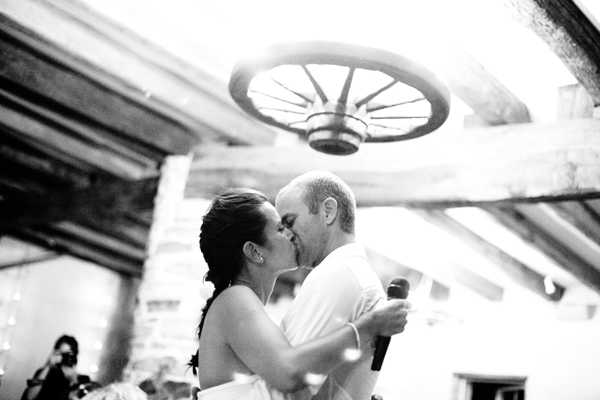 Black-and-white portrait shot of a bride and groom sharing a kiss during what appears to be their first dance or a speech moment at an indoor reception, with the groom holding a microphone. The setting features rustic decor including exposed wooden beams and a decorative wagon wheel mounted overhead, suggesting a barn or farmhouse venue. The bride wears a strapless dress and has her hair in a side braid, while the groom is in a white dress shirt. The image has a high-contrast, slightly overexposed quality with a photographer visible in the soft-focus background.