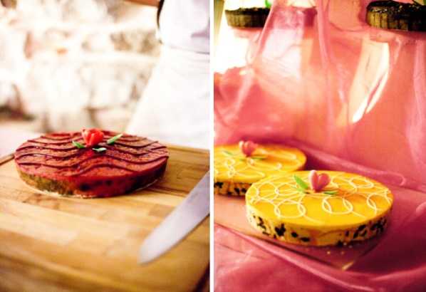 Close-up detail shot of two decorated wedding cakes or tarts displayed side by side in a diptych composition. On the left, a round red-glazed tart with a dark chocolate drizzle pattern and a small red flower garnish sits on a wooden cutting board with a knife beside it, with a person in white attire blurred in the background. On the right, two round yellow cakes decorated with white piped circular patterns and pink flower accents are displayed on bright pink fabric or tissue paper, with a tiered cake stand partially visible in the background. The styling appears colorful and festive with a vibrant, non-traditional palette.