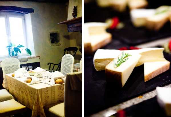 A split image showing two detail shots from what appears to be a wedding reception in a rustic French interior. The left panel shows a small dining table set with a white embroidered tablecloth, wine glasses, and white chair covers in a stone-walled room with a window and fireplace mantel, styled in a classic country French aesthetic. The right panel is a close-up of a cheese course presented on a dark slate board, featuring several wedges of soft-rind cheese garnished with a sprig of rosemary and small red accents, shot with a shallow depth of field.
