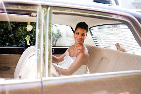 A bride is seated in the back of a vintage wedding car, photographed through the open rear window in a medium portrait shot. She is wearing a strapless white gown and has short dark hair styled close to the head, with a single white flower visible on the car's exterior. The car's interior features cream/ivory upholstered bench seating. She is turned toward the camera with a relaxed expression. The image is taken from outside the vehicle looking in, with greenery visible in the background.