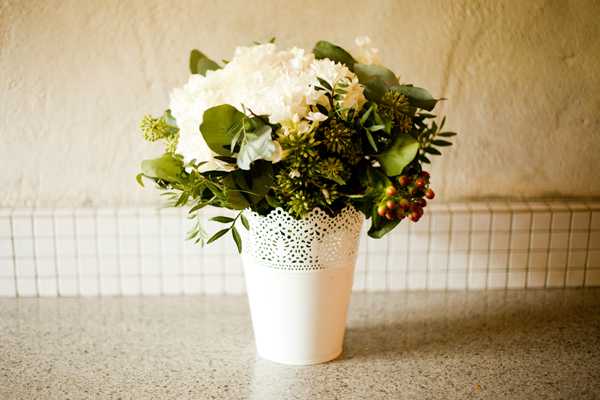 Close-up detail shot of a small floral arrangement sitting on a granite or stone countertop indoors. The arrangement is held in a white metal pot with a decorative laser-cut lace pattern around its upper edge. The flowers include cream hydrangeas, white blooms, green button-like buds, red-green hypericum berries, eucalyptus leaves, and mixed greenery. The overall palette is white, cream, and green with accents of deep red from the berries, suggesting a natural, garden-style floral aesthetic.