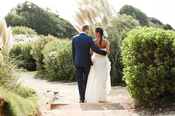 A couple portrait shot from behind showing the bride and groom walking together along a garden path, with the bride's arm around the groom's back. The groom wears a navy blue suit and the bride wears a flowing ivory strapless gown with a long skirt that she is lifting slightly as she walks. The setting is an outdoor garden with trimmed box hedges and tall pampas grass flanking the path. The shot is a medium-distance portrait with a natural, candid feel, taken in daylight with soft, warm lighting.