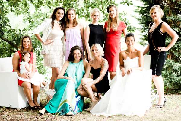 A group portrait of approximately nine women and one small child posing together outdoors in a garden or wooded setting during what appears to be a wedding. The bride is visible near the center-right of the group, wearing a white strapless ball gown. The guests are dressed in a variety of colorful cocktail attire including a red dress, a teal maxi dress, a lavender ruffled dress, a black dress, and dark fitted pants with a black top. One woman seated on a white lounge chair holds a young toddler dressed in red and white. The group is arranged across two rows, with some women standing, some seated, and some crouching at the front. The composition is a wide portrait shot taken in natural daylight with a relaxed, candid posing style.