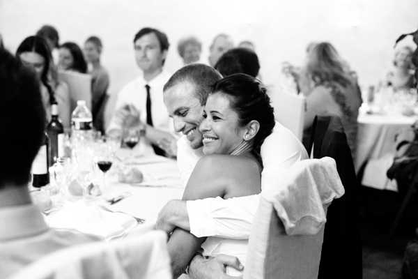 A black-and-white candid shot taken during a wedding reception, capturing the couple seated at the head table embracing and laughing together. The groom has his arms wrapped around the bride, who is wearing an off-the-shoulder dress and has her hair pinned up. The table is set with wine glasses, red wine, water bottles, and place settings with white linens. In the soft-focus background, approximately 8–10 guests are visible standing and seated at nearby tables, dressed in smart-casual attire. The image has strong contrast with bright highlights and mid-range tones, giving it a warm, documentary feel. The composition is a medium portrait shot at table level.