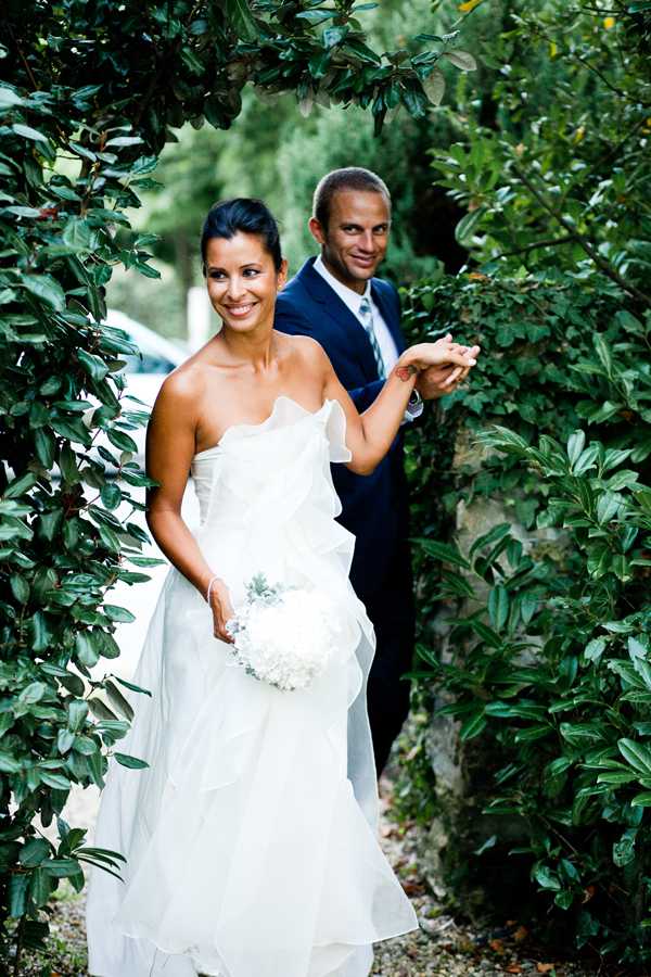 A couple portrait taken outdoors in a garden setting, with the bride and groom framed between dense hedges. The bride wears a strapless white gown with layered, ruffled organza skirt and holds a compact round bouquet of white hydrangeas with touches of dusty miller. The groom stands slightly behind her, wearing a navy blue suit with a light blue striped tie. Both are smiling toward the camera. The shot is a medium portrait with natural daylight, conveying a relaxed, candid mood.