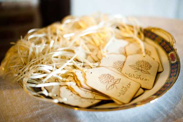 Close-up detail shot of a decorative plate holding a collection of laser-engraved wooden wish tags, each featuring a floral motif and the word 'Wishes' in script, tied with narrow ivory satin ribbons. The tags are arranged loosely on a bed of natural raffia straw within a vintage-style plate with a blue and gold patterned rim. The styling is rustic with natural wood tones, ivory ribbon, and organic textures, likely part of a guest activity station at a reception. The background is softly blurred, showing a linen-covered surface.