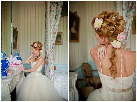 A diptych of getting-ready portraits of a bride inside what appears to be a French chateau room with pale blue-green paneled walls and floral curtains. On the left, the bride stands near a fireplace mantel decorated with blue and white porcelain vases and floral arrangements, adjusting her hair; she wears a full-skirted ivory ballgown. On the right, a close-up rear portrait highlights her upswept hairstyle adorned with blush and pale pink roses, a pearl necklace draped down her back along the lace and button-covered bodice of her gown, and a thin champagne satin sash at the waist. The styling is classic French romantic with vintage-inspired details including the floral hair accessories and pearl embellishments.