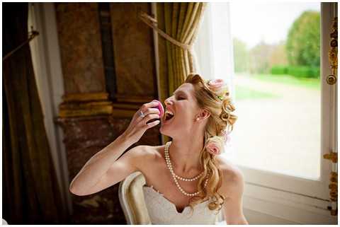 A candid portrait of a bride playfully biting into a pink macaron, laughing with her head tilted back. She is wearing a strapless white lace gown and a pearl necklace, with her blonde hair styled in loose waves accented by small pink roses. The setting appears to be an ornate interior room with gold-toned decorative columns, olive-green drape curtains, and a tall white-framed French door opening onto a green garden beyond. The shot is a medium close-up taken from a slightly elevated angle, capturing the bride's joyful expression mid-bite.