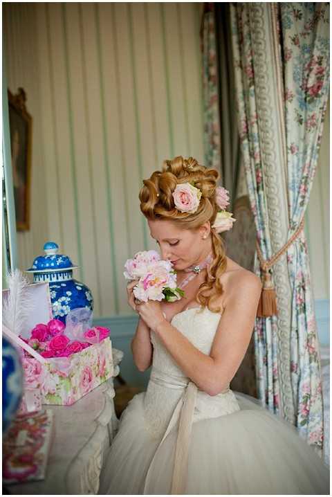 A bride is photographed indoors smelling a pink peony and rose bouquet during a getting-ready moment. She wears a strapless ivory lace ballgown with a satin sash and has an elaborate upswept hairstyle adorned with pink garden roses. The room features floral-print drapes in soft blue and pink tones, striped wallpaper, and a blue-and-white chinoiserie ceramic vase on a side table, alongside a floral-print hat box filled with hot pink roses. The styling has a vintage, Marie Antoinette-inspired aesthetic, and the shot is a medium portrait with soft natural light.