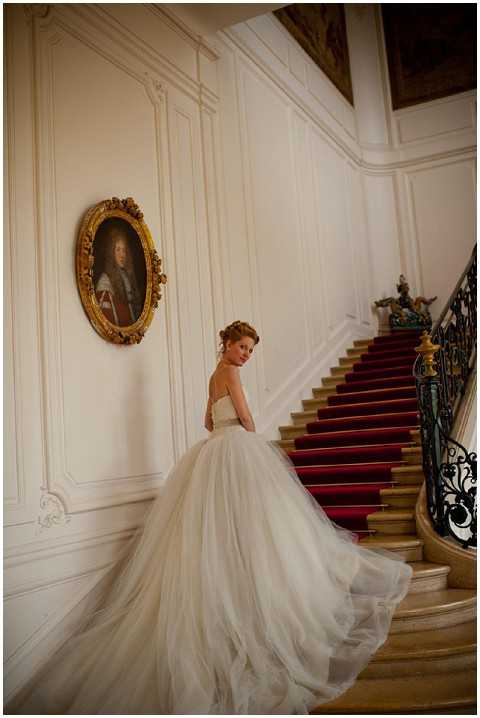 A bridal portrait taken indoors at the base of a grand staircase featuring a deep red carpet runner and ornate black wrought-iron balustrade. The bride stands facing away from the camera and glances back over her shoulder, wearing a strapless ivory ballgown with a voluminous layered tulle skirt and an extended train that fans across the marble floor. Her auburn hair is styled in an updo. The white paneled walls are decorated with a large gilt-framed oval portrait painting in a classic French château style. The composition is a full-length portrait shot taken at a slight angle, emphasizing the dramatic sweep of the dress train against the architectural details of the staircase. Potential venue feature image.