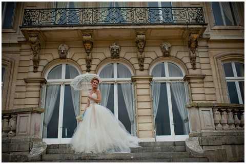 A bride stands alone on the stone steps of a grand French chateau-style building, posed in a portrait shot with a wide angle that emphasizes the ornate limestone facade behind her. She wears a full-skirted ivory ballgown with a cathedral-length train spread across the steps, and holds a white lace parasol. The building features tall arched windows with white curtains, decorative sculpted keystones, wrought-iron balcony railings, and classical carved detailing. The overall styling is classic and formal, consistent with a traditional French estate wedding aesthetic. Potential venue feature image.