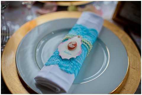 Close-up detail shot of a place setting featuring a white linen napkin rolled and wrapped with a turquoise lace napkin ring, topped with a seashell place card holder displaying a handwritten guest name tag. The napkin rests on a pale grey charger plate with a gold rim, suggesting a coastal or nautical-themed table decor concept. The overall color palette combines turquoise, gold, and soft grey, with the seashell accent reinforcing the beach-inspired styling theme. Additional glassware and table elements are softly blurred in the background.