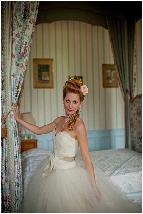 A bridal portrait taken indoors in what appears to be a chateau bedroom during the getting-ready phase. The bride stands in front of a canopy bed, holding back a floral curtain with one hand, and wears an ivory strapless tulle ball gown with a lace bodice and a champagne satin sash at the waist. Her hair is styled in an updo with soft curls, accessorized with a blush pink rose, and she wears a jeweled choker necklace. The room features pale blue wainscoting, green and white striped wallpaper, floral curtains, and gilt-framed artwork on the walls, suggesting a classic French chateau interior with a vintage, romantic decor style. The shot is a medium portrait with soft, warm indoor lighting.