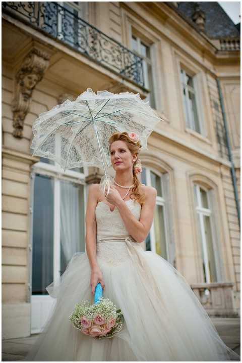 A bridal portrait taken outdoors in front of a classical French stone building with wrought-iron balconies, likely a chateau or hôtel particulier. The bride wears a strapless ivory lace and tulle ballgown with a champagne sash, and accessorizes with a pearl necklace, pearl bracelet, and a small pink flower hair accessory. She holds a white lace parasol in one hand and a bouquet of blush pink roses and white baby's breath with a bright turquoise ribbon wrap in the other. The styling reflects a vintage-inspired, retro aesthetic, and the portrait is shot from a low angle looking up, giving prominence to both the bride and the building facade behind her. Potential venue feature image.