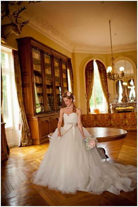 A bridal portrait shot indoors inside what appears to be a chateau library or salon, featuring a bride standing alone on a herringbone parquet wood floor. The bride wears a strapless ivory ballgown with a full tulle skirt, a champagne satin sash at the waist, and a floral hair accessory with a blush pink rose; she holds a round bouquet of soft pink, blush, and white blooms. The room features a large dark wood bookcase filled with books, gold brocade upholstered seating, arched windows with heavy gold drape curtains, and an antique chandelier overhead, suggesting a classic French chateau interior. The composition is a full-length portrait with warm, ambient lighting lending a golden tone to the room.