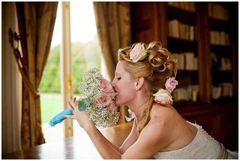 Close-up portrait of a bride smelling her bouquet, composed of blush pink roses and baby's breath with a light blue lace-wrapped stem. She wears a strapless white dress and has an updo hairstyle adorned with blush pink roses. The setting is an indoor room with warm wood bookshelves and tall draped curtains flanking a large window or door that opens to a green outdoor area. The image has a soft, warm tone with shallow depth of field focusing on the bride's face and bouquet.