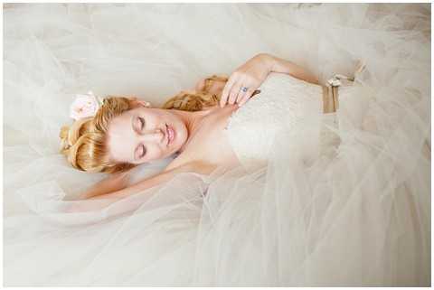 A bridal portrait shot from above showing the bride lying down surrounded by the voluminous tulle skirt of her strapless ivory lace gown, which fans out to fill nearly the entire frame. The bride has wavy auburn hair adorned with a blush pink flower hair accessory, her eyes are gently closed, and she wears a blue gemstone ring on her hand resting near her shoulder. The composition is an overhead close-up portrait with soft, bright, high-key lighting that gives the image a light and airy feel.