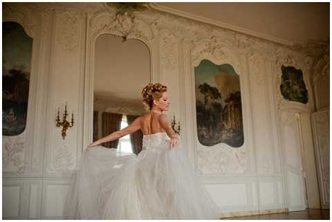 A bride poses indoors in a grand ornate room, caught mid-twirl with the skirt of her strapless white tulle ballgown fanning out around her. She is photographed from behind at a three-quarter angle, her hair styled in an updo with floral or gold hair accessories. The room features elaborate white painted boiserie wall panels with gilded Rococo-style carved molding, antique painted scenic panels depicting pastoral scenes, and a gilt wall sconce with candle arms. The overall interior decor is classic French chateau style. Medium portrait shot with warm ambient lighting. Potential venue feature image.