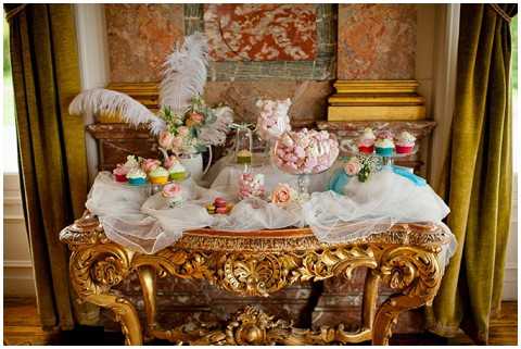 A styled dessert table display set up indoors on an ornate gold gilt Rococo-style console table draped with sheer white fabric. The table holds an assortment of wedding sweets including colorful cupcakes with pastel frosting, pink and white macarons, a glass vessel filled with pink meringues or candy floss, and small floral arrangements featuring blush and coral roses with turquoise accents. Two large white feather plumes rise from a vase on the left side, contributing to the Marie Antoinette-inspired styling theme. The backdrop features gold architectural molding, olive green velvet drapery, and a faded decorative wall panel, suggesting a French chateau interior. Wide shot capturing the full table composition.