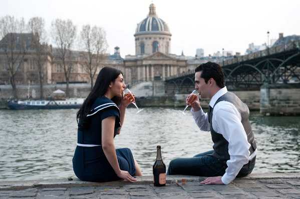 A couple sits facing each other on the stone quayside along the Seine River in Paris, drinking champagne from flutes with a bottle placed between them. The woman wears a navy blue short-sleeved dress with a white sailor-style collar, while the man is dressed in a white dress shirt, grey vest, and dark trousers. The Institut de France with its distinctive gold dome and the Pont des Arts bridge are visible in the background across the river, placing the shoot in central Paris. This is an outdoor engagement or couple portrait session captured as a medium wide shot with a relaxed, candid feel.