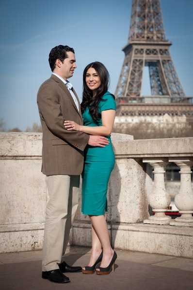 A couple poses together on a stone balustrade terrace with the Eiffel Tower prominently visible in the soft-focus background, suggesting an engagement or pre-wedding portrait session in Paris. The man wears a tan blazer, khaki trousers, and a light blue tie, while the woman wears a fitted teal/turquoise knee-length dress with black platform heels and long dark wavy hair. They are standing close together, his arm around her waist and her hand resting on his chest, both smiling. The shot is a full-length portrait taken at street level with a clean, bright exposure.