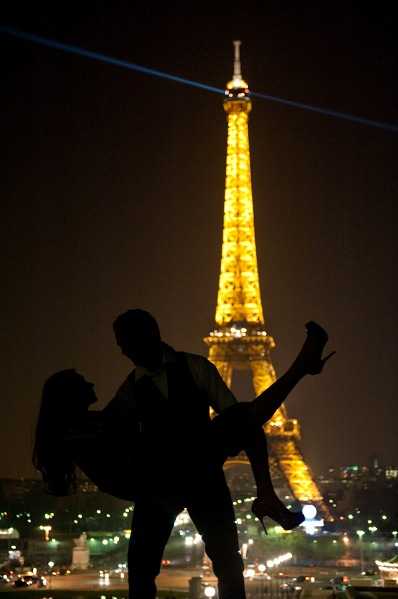 A couple portrait taken at night in Paris, with the Eiffel Tower fully illuminated in gold and its rotating searchlight beam visible against the dark sky. The couple appears as a complete silhouette in the foreground — the groom lifting the bride, whose legs are raised and high heels are visible — creating a dramatic contrast against the brightly lit tower. The city lights of Paris are visible across the background at street level. Wide shot with a centered composition emphasizing the iconic landmark behind the couple.
