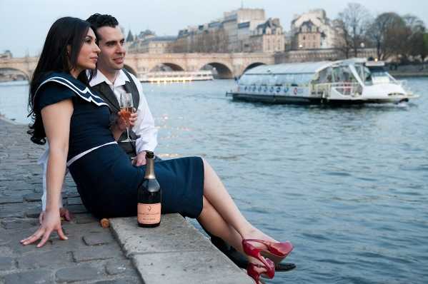 A couple sits together on the stone quayside of the Seine River in Paris during what appears to be an engagement or pre-wedding portrait session. The woman wears a fitted navy blue dress with a white sailor-style collar and bright red heels, while the man wears a white dress shirt and dark trousers; both hold champagne flutes and a bottle of rosé champagne rests on the ledge beside them. They are gazing toward the river where a bateau mouche and the Pont Neuf bridge are visible in the background, giving the scene a distinctly Parisian urban setting. The shot is a medium-distance portrait taken at quay level with soft overcast light, and the overall styling has a retro-nautical feel with the coordinated navy and red color palette.
