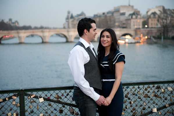 A couple poses for an engagement or pre-wedding portrait on the Pont des Arts in Paris, with the Seine River and Pont Neuf visible in the background at dusk. The man wears a white dress shirt, grey vest, and dark tie, while the woman wears a navy blue short-sleeve dress with white trim detail. The railing in the foreground is lined with numerous padlocks, characteristic of this Paris bridge. The shot is a medium portrait taken at eye level, with city lights beginning to glow softly in the blue-hour background.