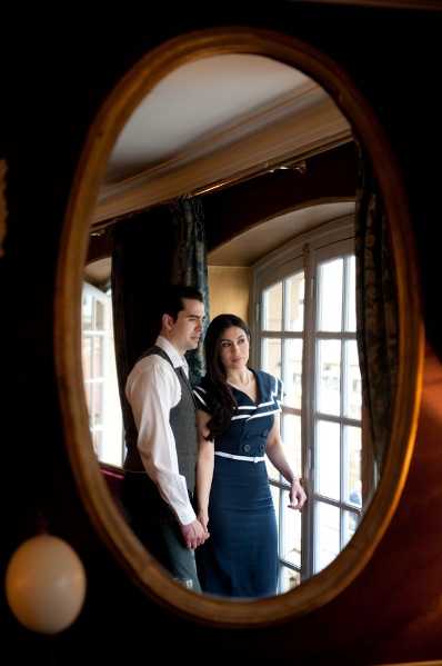 A couple is reflected in an oval gold-framed mirror inside what appears to be a classic French interior with large multi-pane windows and dark curtains. The groom wears a white dress shirt with a grey waistcoat and dark trousers, while the bride wears a fitted navy blue dress with white contrast detailing, giving the overall styling a vintage 1940s-inspired aesthetic. The couple stands close together, facing the camera with serious expressions, with bright window light backlighting them from behind. The shot is a creative portrait composed entirely within the oval mirror reflection, with the dark foreground framing adding depth and contrast.