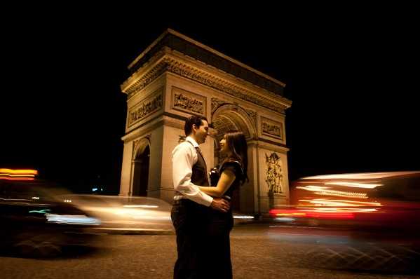 A couple poses facing each other in an intimate portrait in front of the Arc de Triomphe in Paris at night. The man wears a white dress shirt with a dark tie and dark trousers, while the woman wears a dark dress. The shot uses a slow shutter speed, creating motion blur from passing vehicle headlights and taillights that form streaks of warm orange and red light on either side of the couple, who remain sharp against the illuminated monument. This appears to be an engagement or pre-wedding portrait session rather than a wedding day shoot. The wide shot frames the couple centrally with the Arc de Triomphe lit up directly behind them against a black night sky.