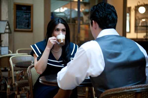 A couple sits together at a small bistro table inside what appears to be a Parisian-style café, in a relaxed engagement or pre-wedding portrait session. The woman, facing the camera, wears a navy blue and white short-sleeve dress with a pearl necklace and sips from a glass coffee cup containing a layered latte. The man, shown from behind, wears a white dress shirt with a grey vest. The café setting features wicker bistro chairs, a chalkboard menu on the wall, and warm ambient lighting visible in the background. Medium close-up portrait shot with a shallow depth of field.