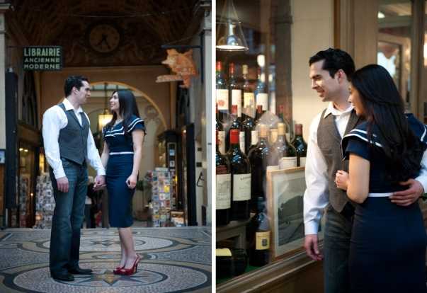 A couples engagement or pre-wedding portrait session shown in two side-by-side images taken inside a Parisian covered arcade (galerie), with a sign reading 'Librairie Ancienne Moderne' visible in the background of the left frame. The man wears a white dress shirt with a grey herringbone vest and dark jeans, while the woman wears a fitted navy blue dress with white trim detailing and red heels. In the left image, the couple stands facing each other holding hands on the ornate tiled floor of the arcade, captured in a full-length wide portrait. The right image is a closer portrait showing the couple embracing in front of a wine shop display with multiple wine bottles visible behind glass, lit by warm pendant lighting overhead. The styling has a classic, vintage-inspired Parisian aesthetic.