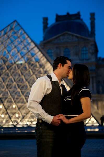 A couple shares a kiss during an outdoor evening portrait session in front of the illuminated glass pyramid at the Louvre in Paris, with the historic museum building visible in the background. The man wears a white dress shirt with a dark grey vest and tie, while the woman wears a dark navy short-sleeved top with white striped detailing. The shot is taken during blue hour, with the pyramid's internal lighting creating a warm glow against the deep blue twilight sky. The image is a medium full-length portrait with the architectural landmark centered behind the couple.