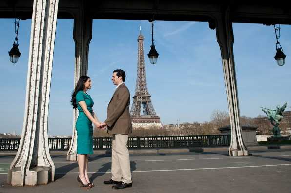 A couple stands facing each other and holding hands on the Bir-Hakeim bridge in Paris, with the Eiffel Tower prominently visible in the background. The woman wears a fitted teal/emerald green knee-length dress with dark hair worn down, while the man wears a brown blazer with khaki trousers. The shot is framed by the ornate iron columns and hanging lanterns of the bridge's elevated metro structure. This appears to be an engagement or pre-wedding portrait session, captured as a medium full-body shot in natural daylight.