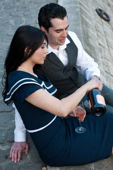 A couple sits together on stone steps in what appears to be an outdoor portrait session, likely an engagement or pre-wedding shoot. The woman wears a navy blue dress with white sailor-collar detailing and a pearl necklace, while the man is dressed in a white shirt and grey tweed vest. She is pouring rosé wine from a large bottle into two stemmed glasses, and both are looking down at the pour. The shot is a medium close-up portrait with a shallow depth of field, slightly blurring the stone background.