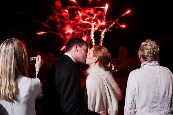 A couple kisses outdoors at night during a fireworks display, with large red and orange fireworks bursting in the dark sky directly behind them. The groom wears a dark suit and the bride wears a cream or ivory wrap or shawl with white fur trim over her wedding attire, with her auburn hair styled up. Two female guests are visible on either side — one on the left photographing the moment with a small camera or phone, and one on the right with an updo hairstyle wearing a light linen jacket. The shot is a close portrait-style image taken from behind and slightly to the side, with the fireworks serving as a dramatic backlit backdrop.