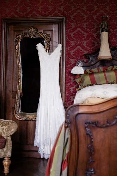 A getting-ready detail shot of a white lace wedding dress hanging from an ornate gold-framed floor mirror in a chateau-style bedroom. The dress features a V-neckline, delicate lace detailing, and a flowing A-line silhouette. The room is decorated with deep red damask wallpaper, a carved dark wood bed with striped and floral pillows, a wall sconce, and an antique upholstered chair visible at the left edge, creating a classic French interior setting. The composition is a medium wide shot emphasizing the dress against the richly decorated baroque-style room.