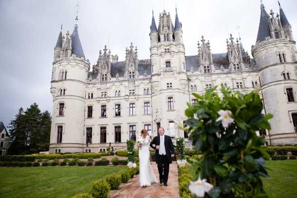 A bride and groom walk together along a garden path in front of a large white French chateau with dark slate-roofed turrets and ornate Gothic-Renaissance architecture. The bride wears a long-sleeved, fitted white lace gown and carries a bridal bouquet, while the groom is dressed in a dark suit with a white shirt. The shot is taken from a medium-wide angle at ground level, with a flowering shrub with white blooms in the soft foreground creating natural depth. The overcast sky and manicured box hedges frame the couple as they move toward the camera. Potential venue feature image.
