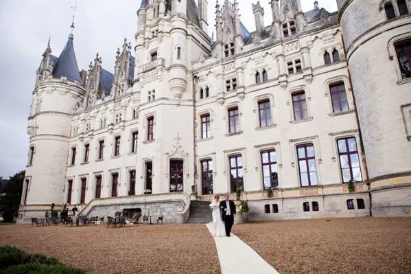 A bride in a white gown and groom in a dark suit stand together on a white-paved pathway in the gravel courtyard of a large French Renaissance-style chateau, photographed as a wide shot that emphasizes the scale of the white stone building with its ornate towers, spires, and Gothic detailing. A small group of guests or staff is visible near the entrance steps to the left, where outdoor furniture — black chairs and tables — is arranged on the courtyard. The image is taken in overcast natural light, giving the scene a muted, even tone. Potential venue feature image.