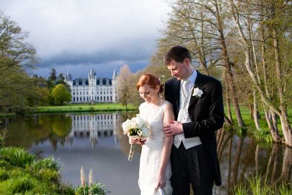 A couple portrait taken outdoors on the grounds of a French chateau, with the white multi-story chateau building reflected in a large ornamental lake in the background. The bride has short red hair and wears a sleeveless white fitted gown, holding a compact bouquet of white flowers including what appear to be peonies or ranunculus. The groom wears a black morning coat with grey striped trousers, a white waistcoat, and a white boutonniere. He stands behind the bride with his arm around her as both look downward. The styling is classic and formal. The shot is a medium portrait framed horizontally, with the lake and chateau providing a distant backdrop. Potential venue feature image.