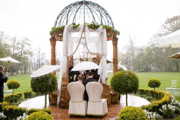 An outdoor wedding ceremony taking place beneath an ornate wrought-iron dome gazebo with wooden columns, draped with white fabric and decorated with green foliage along the top. The couple is seated in large cream upholstered high-back chairs on a circular tiled platform, with an officiant and guests holding white parasols to shelter from visible rain. The formal garden setting features manicured round topiary shrubs, white flowering plants, and a well-kept lawn extending into the background. The overall decor style is classic and formal, with a cream and white palette; the image is taken as a wide shot from behind the couple, showing the full gazebo structure and surrounding garden layout. Potential venue feature image.