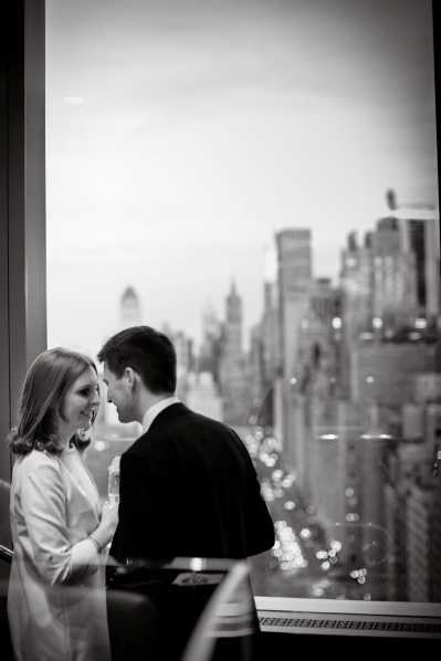 A black-and-white portrait of a couple standing close together and facing each other in front of a large floor-to-ceiling window, with a sweeping urban skyline visible in the background suggesting a high-rise venue in a major city. The woman wears a light-colored jacket or blazer and smiles up at the man, who is dressed in a dark suit. Bokeh city lights are visible along the street below, adding depth to the background. The image is a medium close-up shot with a slightly soft focus, taken from a low angle with strong contrast between the couple in the foreground and the bright window behind them.