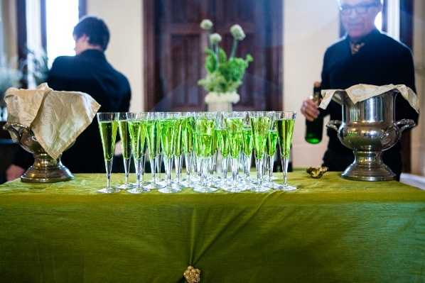 A cocktail hour drinks station set up indoors, featuring approximately 20–25 champagne flutes filled with a bright green liquid arranged in rows on a table draped with an olive green cloth. Two staff members dressed in black are visible in the background — one holding a dark green bottle and standing near a silver ice bucket on the right, and another with their back turned on the left. A floral arrangement of green globe thistle or similar round green blooms in a white vase is visible in the background, reinforcing the all-green color palette. The shot is taken from a low angle at table level, giving a close-up detail perspective of the glassware display.