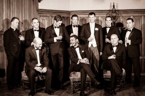 A sepia-toned group portrait of the groom and approximately nine groomsmen posed in a richly wood-paneled interior room, likely inside a chateau or manor house. The men are arranged in two rows — four seated on ornate carved wooden chairs in the foreground and six standing behind — all wearing black tuxedos with bow ties and white dress shirts, with the groom distinguished by a white waistcoat and boutonniere. Several of the men are holding champagne flutes and laughing candidly, giving the image an relaxed, celebratory feel. The shot is a wide group portrait with warm sepia tones that complement the dark wood paneling and parquet flooring of the formal interior setting.