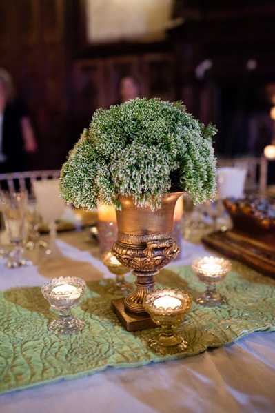 Close-up detail shot of a wedding reception table centerpiece set in a dimly lit indoor venue, likely a historic chateau or manor hall. The centerpiece features a dense arrangement of green sedum or similar textural greenery in an ornate antique gold urn with decorative relief detailing. The table is dressed with a white linen base and a sage green embroidered or quilted table runner, with several small crystal and gold votive candle holders casting warm candlelight around the centerpiece. The overall decor palette is gold, sage green, and ivory, suggesting a classic, old-world styling theme with an emphasis on rich metallics and lush greenery rather than traditional floral blooms.