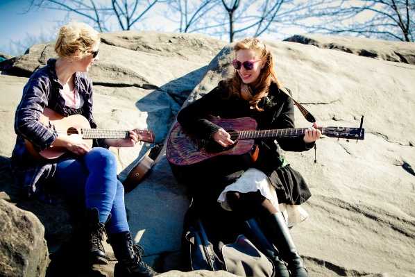 This image does not appear to be a wedding photograph. It shows two young women sitting on large rocks outdoors, playing stringed instruments — one holds a small ukulele and the other plays an acoustic guitar with a decorative reddish finish. Both are dressed in casual everyday clothing including jeans, dark jackets, and boots, with no visible wedding attire, floral arrangements, or wedding-related decor. This image does not appear suitable for a French wedding content library.