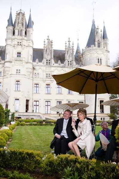 Wedding guests are seated outdoors in a formal garden during what appears to be a cocktail hour or outdoor ceremony, with a large yellow market umbrella and a smaller ivory parasol providing shade. In the foreground, a man in a dark suit and a woman in a white/cream coat sit together conversing, with at least one other guest partially visible to the right. The setting is the formal garden grounds of a grand French Loire Valley-style chateau featuring multiple pointed slate turrets, ornate Gothic-Renaissance architecture, and a white stone facade directly behind the guests. The shot is a medium wide portrait framing the guests with the chateau prominently filling the background. Potential venue feature image.