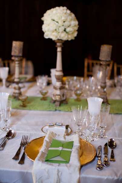 Close-up detail shot of a wedding reception table setting indoors against a dark background. The place setting features a gold charger plate topped with a folded ivory linen napkin and a green folded menu card or place card holder secured with a silver buckle detail. Multiple crystal glasses are arranged to the right, and silver flatware is placed on both sides. A green table runner runs down the center of the white linen tablecloth. In the background, a tall white pedestal stand holds a dense pomander-style centerpiece of white roses, flanked by silver candle holders with pillar candles and additional crystal glassware, suggesting a classic, formal reception decor palette of white, gold, green, and silver.